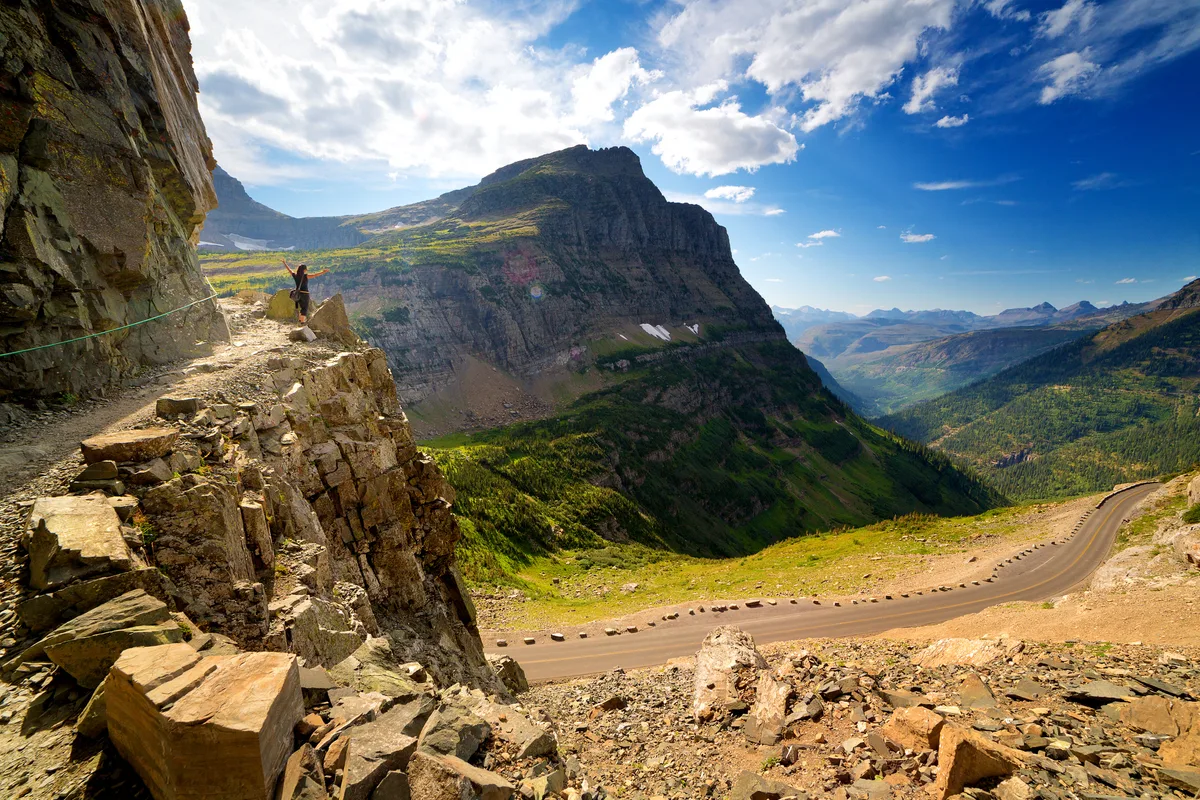 Female hiker on narrow highline trail stands on overlook with arms outstretched, looking down on going-to-the-sun road not far from Logan pass in glacier National Park, Montana, on partly cloudy summer afternoon.